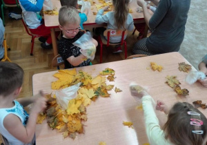Dzieci siedzą przy stolikach, wkładają liście do torebeczek