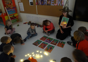 Nauczycielka pokazuje dzieciom ilustracje dotyczące tradycji andrzejkowych.