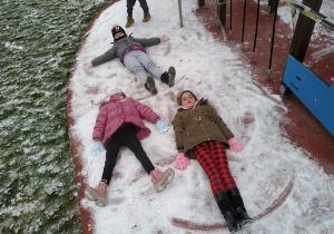 Zuzia, Malika i Oliwia robią anioła na śniegu.