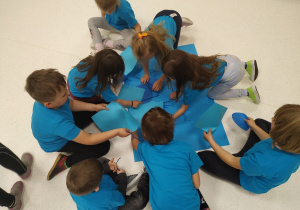 Dzieci ubrane w niebieskie koszulki siedzą na podłodze i sięgają po niebieskie kartki.