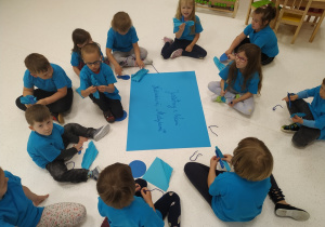 Dzieci ubrane w niebieskie koszulki siedzą na podłodze i wykonują pracę plastyczną.