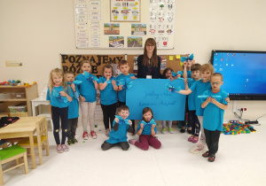 Dzieci ubrane w niebieskie koszulki pozują do zdjęcia. Dzieci trzymają zrobione przez siebie motyle. Pani krzyma niebieski karton z hasłem "Jesteśmy z Wami Niebieskimi Motylami"