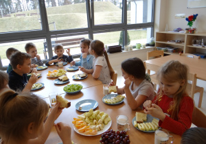 Dzieci siedzą przy stole i zajadają się urodzinowymi owocami.