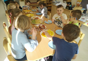 Dzieci siedzą przy stole i zjadają urodzinowe owoce.