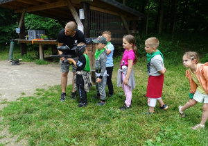 Igorek celuje do tarczy z pistoletu do paintballa. Pozostałe dzieci czekają w kolejce i przyglądają się.