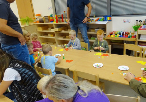 Dzieci z rodzicami siedzą przy stolikach wykonują pracę plastyczną - wyklejają kolorowymi kółkami motyle.