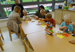 Dzieci z rodzicami siedzą przy stolikach i wykonują pracę plastyczną.