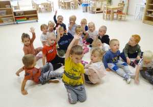 Dzieci siedzą na podłodze podnosząc ręce do góry, kiedy mowa jest o czymś, co lubią robić.