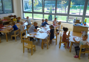 Maluszki siedzą przy stolikach. Każde ma kartkę oraz pędzelek i malują farbami. Niektóre dzieci maczają pędzelek w farbie znajdującej się na podstawku.