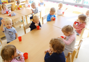 Dzieci siedzą przy stolikach i jedzą galaretki z czerwonych kubeczków.