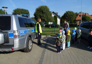 Dzieci wraz z panią stoją na przedszkolnym parkingu obok radiowozu i słuchają pani policjantki.