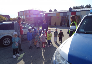 Maluszki z paniami stoją na parkingu i słuchają opowieści pani policjantki.