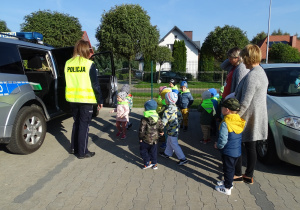 Pani policjantka otworzyła drzwi radiowozu i zaprasza do niego dzieci.