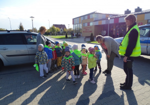 Maluchy stoją z paniami i panią policjantką na parkingu przedszkola, na szyjach mają apaszki odblaskowe, które otrzymały od pani gościa.