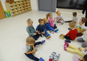 Dzieci siedzą na podłodze, w rękach trzymają male karty pracy z klamerkami.