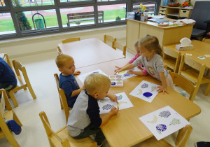 Dzieci siedzą przy stole i wykonują pracę plastyczną metodą stemplowania.