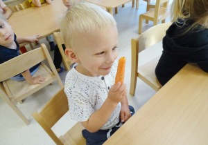 Uśmiechnięty Ernest siedzi przy stoliku, trzyma marchewkę i wącha ją.