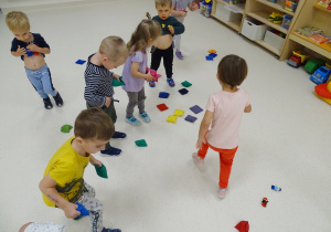 Dzieci zbierają porozrzucane po podłodze kolorowe woreczki gimnastyczne.