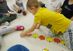 Dzieci siedzą na podłodze. Ksawery kładzie żółte jabłuszko, aby uzupełnić sekwencję z czerwonych i żółtych jabłek.