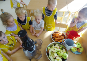 Dzieci stoją wokół stolika, na którym postawione są miski z jabłkami, marchewkami oraz wyciskarką do owoców.