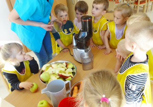 Dzieci stoją wokół stolika, na którym postawione są miski z jabłkami, marchewkami oraz wyciskarką do owoców. Pani tłumaczy dzieciom zasady obowiązujące podczas przygotowywania soku.