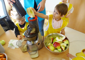 Blanka wrzuca jabłko na sok, pani jej pomaga. Pozostałe dzieci obserwują.