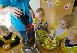 Kostek wrzuca jabłko na sok, pani mu pomaga. Pozostałe dzieci obserwują.