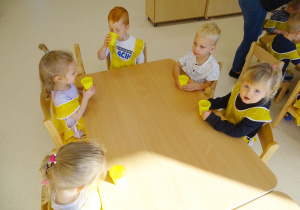 Dzieci siedzą przy stoliku i próbują sok z żółtych kubeczków.