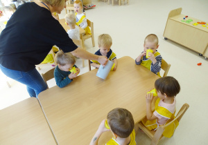Dzieci siedzą przy stoliku i piją sok z żółtych kubeczków. Pani dolewa im soku z dzbanka.