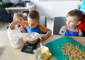 Leon przesypuje łyżką mąkę do miski znajdującej się na wadze. Pozostałe dzieci się przyglądają.