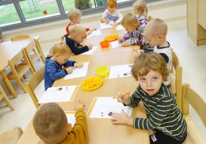 Dzieci siedzą przy stolikach i kolorują wiewiórki kredką świecową.