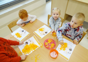 Dzieci siedzą przy stoliku i wyklejają ogon wiewiórki pomarańczowymi ścinkami.