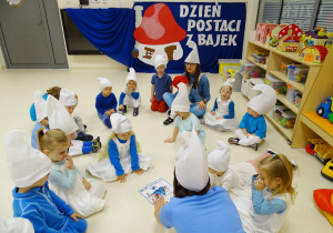 Dzieci siedzą w kole. Anielka i Ksawcio układają z pomocą pani puzzle Smerfa Ważniaka.