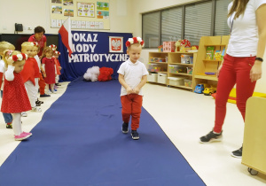 Kostek ubrany na biało czerwono pozuje podczas "Pokazu mody patriotycznej" na granatowym dywanie.