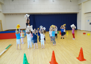 Dzieci i pani ćwiczą z misiami na sali gimnastycznej podnosząc ręce wysoko do góry.