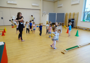 Dzieci i pani ćwiczą na sali gimnastycznej przekładając misie na boki.