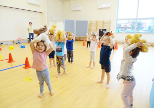 Dzieci stoją na sali gimnastycznej, trzymając misie na głowach.