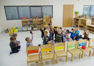 Dzieci siedzą na podłodze. Trzymają ręce w górze i pokazują węgla wykonane z papieru. Przed nimi stoją krzesełka z kolorowymi kartkami.