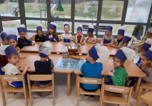 Dzieci w strojach kucharzy siedzą wspólni wokół złączonych stolików