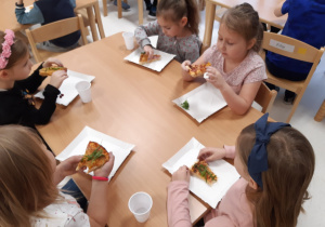 Dzieci siedzą przy stoliczkach i zajadają się pizzą