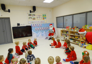 Maluchy siedzą na podłodze, przed nimi na krześle siedzi święty Mikołaj, który pokazuje im grę.