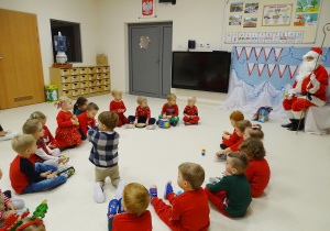 Maluchy siedzą na podłodze, trzymają instrumenty muzyczne, przed nimi na krześle siedzi święty Mikołaj.