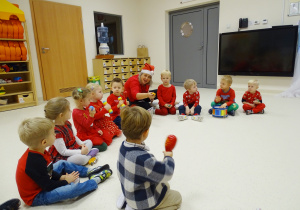 Maluchy siedzą na podłodze i razem z panią Olą śpiewają oraz grają dla świętego Mikołaja na instrumentach.