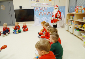 Maluchy siedzą na podłodze i razem z panią Olą śpiewają oraz grają dla świętego Mikołaja na instrumentach.