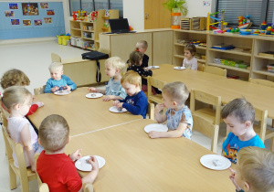Dzieci siedzą przy stolikach i jedzą wyłożone na talerzykach ciasteczka.