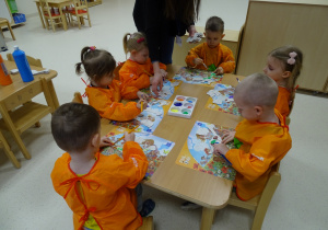 Dzieci siedzą przy stolikach w fartuszkach malarskich i malują farbami.