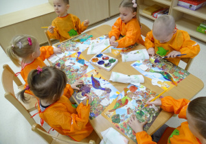 Dzieci siedzą przy stolikach w fartuszkach malarskich i malują farbami model dinozaura.