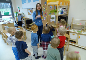 Dzieci stoją z nauczycielką i mrówką - gościem przy obrazku z hipopotamem i słuchają opowiadania o nim.