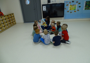 Dzieci siedzą w kole i obserwują obrazki, które pokazuje im nauczycielka.