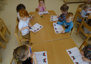 Dzieci siedzą przy stoliku i wycinają kartkę nożyczkami wzdłuż linii.